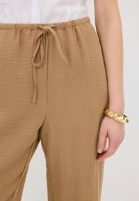Bruine, gestructureerde, brede broek met een elastische tailleband en een trekkkoord. Een gouden kettingarmband siert de pols van de getoonde hand.