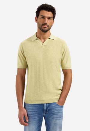 Man met kort krullend haar en baard, gekleed in een lichtgeel poloshirt met korte mouwen en blauwe spijkerbroek, staand met één hand in zijn zak.