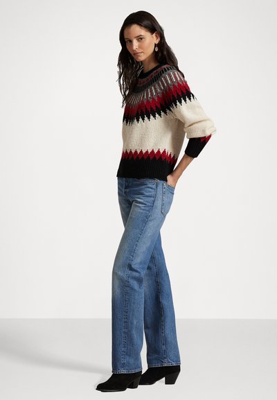 Kötött pulóver krém alappal, amely piros, fekete és ezüst geometrikus mintákkal díszített, világoskék, egyenes szárú farmernadrággal és fekete bokacsizmával párosítva.