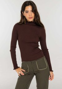 Top marrón de manga larga acanalado con cuello alto tipo mock y bordes con volantes, combinado con pantalones de cuadros verdes y negros con ribetes contrastantes.