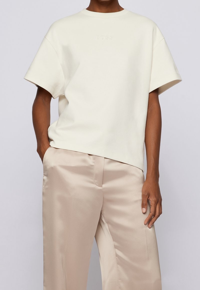 Personne portant une chemise blanche ample à manches courtes avec un logo subtil "BOSS" et un pantalon en satin beige clair, les mains dans les poches.