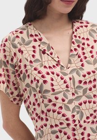 Blusa a fiori beige con fiori rosa e rossi, foglie grigie, maniche corte e scollatura a v. Tessuto dalla texture liscia.