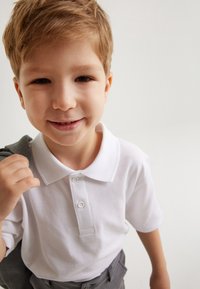 Camisa polo blanca con cuello y frente de botones, usada por un niño, combinada con pantalones grises, sonriendo mientras sostiene una chaqueta gris.