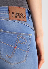 Light blue denim jeans with a leather patch labeled "PINKO JEAN" and distinctive stitching detail featuring a stylized "P" and "J."