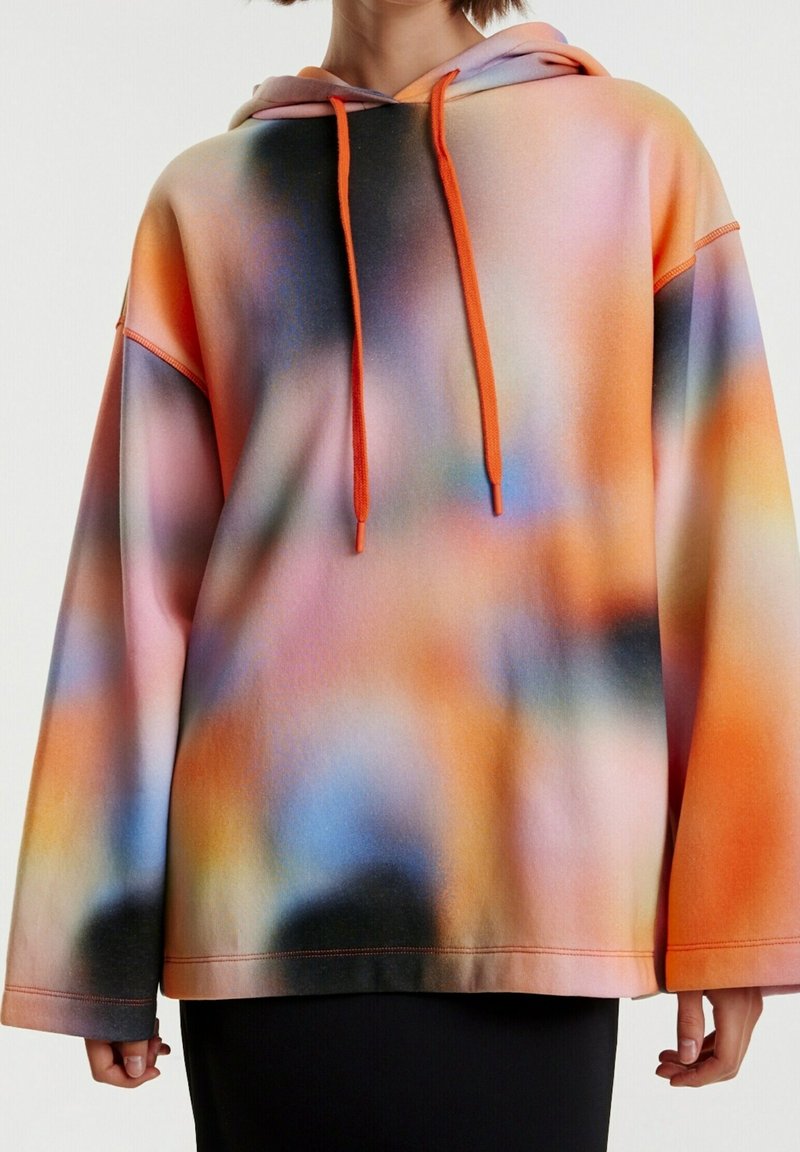 Sweat à capuche multicolore à effet flou doux avec des cordons orange porté sur une jupe noire, montrant le torse et les bras sur un fond uni.