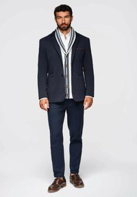 Blazer azul marino con solapas en contraste, camisa blanca con una bufanda de rayas, pantalones oscuros, zapatos marrones de cordones y botones en el cárdigan.