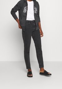 Grijze ritshoodie met wit logo, gecombineerd met zwarte slim-fit jeans en zwarte instapsandalen. Glad materiaal, comfortabele pasvorm en minimalistisch ontwerp.