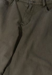 Nahaufnahme von Stoff einer dunkelolivgrünen Hose, die den Bund, Knopf, Reißverschluss, Gürtelschlaufen und Details der vorderen Taschennaht zeigt.