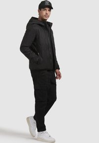Schwarze Pufferjacke mit hexagonalem gestepptem Design, mit Reißverschluss an der Vorderseite, Kapuze, kombiniert mit schwarzen Cargo-Hosen und weißen Sportschuhen.