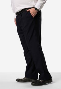 Schwarze, maßgeschneiderte Hosen mit geradem Bein, seitlichen Taschen und glatter Textur, kombiniert mit schwarzen Schuhen. Weißes Hemd und Uhr sind sichtbar.