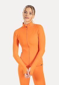 Orange atletisk zip-up jacka i stretchigt material, med hög krage och hål för tummen vid ärmsluten, kombinerad med matchande leggings.