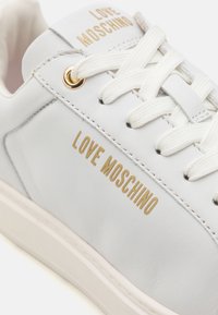 Biele kožené tenisky s zlatým brandingom "LOVE MOSCHINO", ploché biele šnúrky a béžová gumová podrážka. Hladká textúra, minimalistický dizajn.