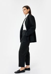 Zwarte op maat gemaakte blazer over een witte coltrui, gecombineerd met zwarte broek en loafers. Stof lijkt glad; het jasje heeft gestructureerde schouders.