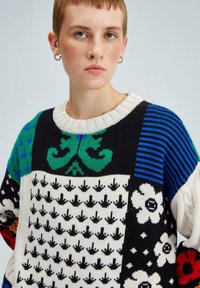 Pull en tricot multicolore présentant divers motifs : fleurs vertes, flèches noires, rayures bleues et fleurs blanches, avec un col rond côtelé.