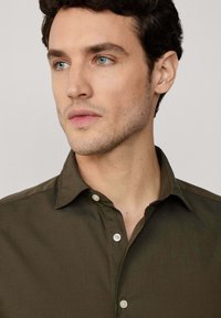 Camisa verde oliva oscura con una textura suave, cuello abotonado y botones blancos. Presenta un diseño ajustado y detalles de estilo minimalista.