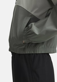 Lehká olivově zelená bunda s kontrastním tmavším panelem, kapsou na zip a elastickými manžetami a lemem. Vhodná pro venkovní nošení.