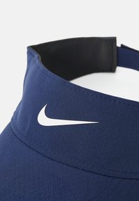 Marineblå skyggehatt laget av stoff med teksturert finish; har en hvit Nike-logo på fronten, og et svart indre bånd.