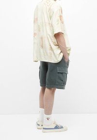 Camicetta beige chiaro a maniche corte con testo colorato e motivi floreali, abbinata a pantaloncini cargo verdi e scarpe da ginnastica con dettagli blu e calze bianche.