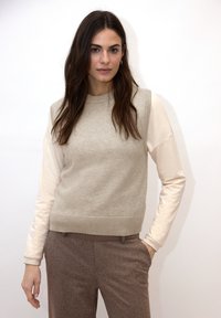 Pull en maille beige avec un ourlet côtel é et des épaules côtelées, associé à une chemise à manches longues beige clair en dessous, porté avec un pantalon marron texturé.
