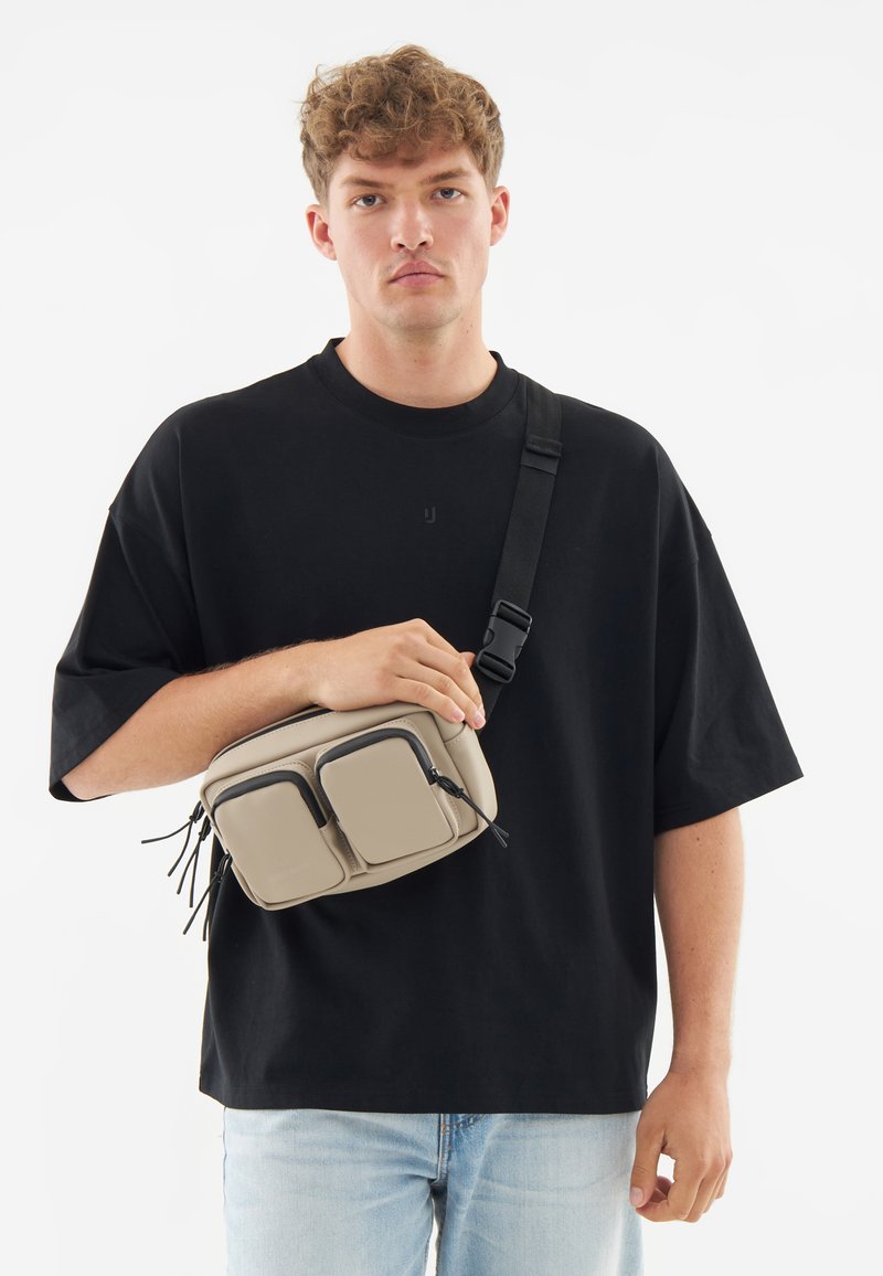 Beige axelremsväska med två framfickor, svarta dragkedjor och kvastdetaljer. Bärs av en person klädd i en svart oversized t-shirt.