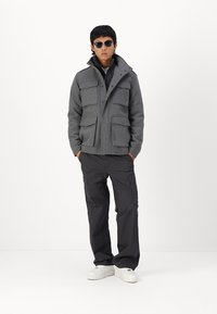 Chaqueta de mezcla de lana gris con cuatro bolsillos exteriores, cremallera frontal y cuello; combinada con pantalones cargo negros y zapatillas blancas.