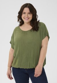 Mujer sonriente con cabello castaño ondulado hasta los hombros, vistiendo una blusa de manga corta verde oliva y unos jeans oscuros, frente a un fondo plano.