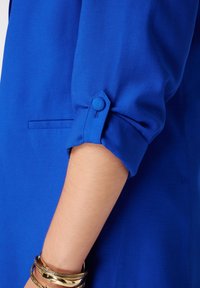 Tissu de chemise bleu au toucher doux, avec des manches retroussées maintenues par des détails de bouton. Bracelets en or au poignet.