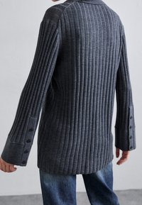 Person, der einen dunkelgrauen gerippten Pullover mit langen Ärmeln trägt, der über Knopfmanschetten verfügt, kombiniert mit blauen Jeans, von hinten gezeigt.