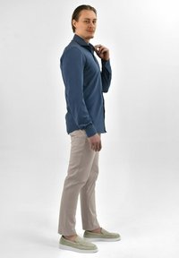 Donkerblauwe overhemd met lange mouwen en knopen, gecombineerd met beige slim-fit broeken en lichtgroene instappers, staande tegen een effen achtergrond.