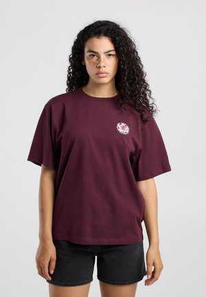 Jonge vrouw met krullend haar die een donkerrood t-shirt draagt met een klein rond bloemenlogo en zwarte korte broek, staand tegen een effen achtergrond.