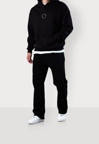 Mężczyzna w czarnej bluzie z kapturem z białym okrągłym napisem, czarnych spodniach, białych trampkach, stojący na tle jasnoszarego tła.