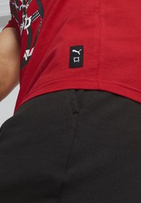 Červené tričko s krátkým rukávem a grafickým designem, na lemu s černou značkovou nášivkou Puma. Spárováno s tmavě šedými kalhotami.