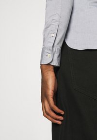 Camicia di cotone azzurro chiaro con maniche lunghe, polsini abbottonati e orlo curvo. Primo piano di una mano appoggiata su uno sfondo di tessuto scuro.