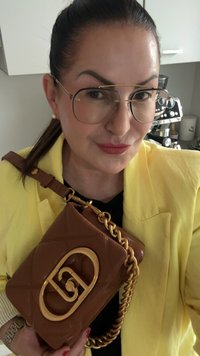 Bolso acolchado marrón con herrajes dorados, que presenta un logo distintivo y una correa de cadena, sostenido contra un blazer amarillo y una blusa negra.