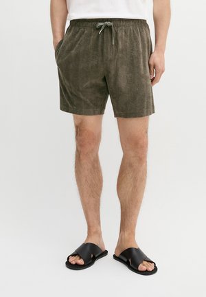 Man draagt olijfgroene, gestructureerde shorts met trekkoord en zwarte slides met gekruiste bandjes, handen in de zakken, tegen een effen achtergrond.