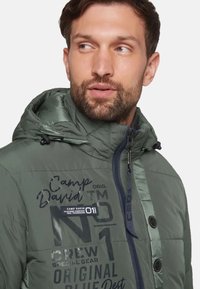 Grön pufferjacka med huva, quiltat mönster och svarta detaljer. Har branding och textmönster på bröstet samt en dragkedjestängning.