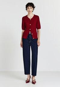 Donna in cardigan rosso a maglia grossa a maniche corte, jeans dritti a vita alta blu scuro e tacchi a punta rossi coordinati su sfondo neutro.