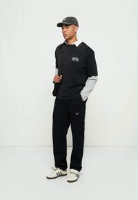 Juoda ilgomis rankovėmis marškinėliai su pilkomis rankovėmis ir logotipu, juodos kelnės su kišenėmis ir pilka kepurė, užbaigti baltais ir pilkais sportbačiais.