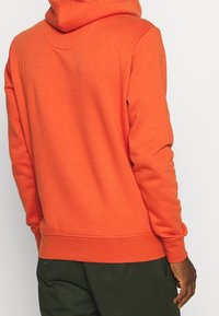 Orange huva sweatshirt med en texturerad tyg. Har en känguruficka, ribbade muddar och nederkant. Synlig sömnad längs halsringningen.
