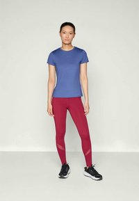 Femme debout portant un t-shirt de sport bleu, des leggings rouges avec des détails en mesh et des baskets noires, sur un fond clair uni.