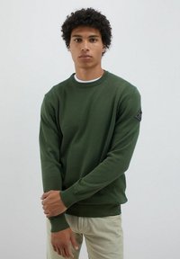 Maglione verde lavorato a maglia con scollatura rotonda e polsini a costine. Presenta un badge nero sulla manica sinistra. Indossato sopra una camicia bianca.