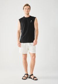 Chemise sans manches noire en coton avec un petit logo de palmier, associée à un short texturé blanc et des sandales noires.