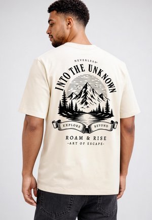 Man wearing beige t-shirt with black mountain and forest graphic and text "Into the Unknown, Explore Beyond, Roam & Rise" on the back.
