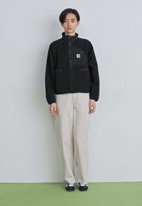 Joven de pie, vistiendo una chaqueta negra con cremallera, pantalones beige y zapatillas en blanco y negro, contra un fondo gris claro liso.