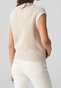 Ljusbeige stickad ärmlös tröja med ribbad nederkant, med texturerad tyg och figurnära design, kombinerad med vita tajta jeans.