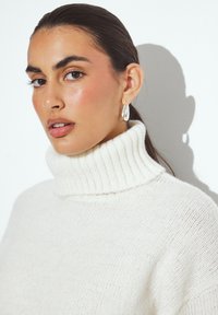 Baltas megztinis su aukštu kaklu ir raukšlėta tekstūra. Modelis turi minimalų makiažą, matomos sidabrinės auskarai žiedais, o fone yra neutralus fonas.