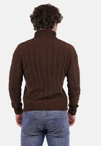 Maglione marrone a trecce con collo alto, motivo testurizzato e polsini a costine. Indossato con jeans azzurri, visto da dietro.