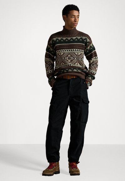 Sweater de malha com um padrão geométrico em castanho, verde e creme, combinado com calças cargo pretas e botas de atacadores castanhas com detalhes em vermelho.