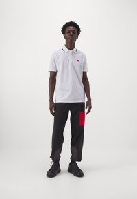 Polo bianco con colletto nero e logo rosso, abbinato a pantaloni neri con dettagli rossi e motivo decorativo. Sneakers nere.