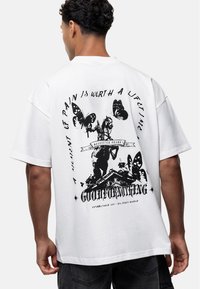 Biała koszulka z krótkim rękawem z grafiką konia i motyli w czarnym nadruku, z tekstem i dekoracyjnymi elementami z tyłu.